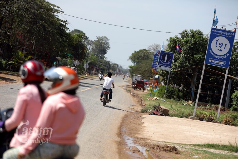 ប្រជាពលរដ្ឋធ្វើដំណើរឆ្លងកាត់ផ្លូវមួយដែលមានស្លាកគណបក្សខ្មែរស្រឡាញ់ជាតិ និង គណបក្សភ្លើងទៀន ដាក់តាំងបង្ហាញ ក្នុងខេត្តកណ្ដាល។ រូបថតថ្ងៃទី៨ ខែមិនា ឆ្នាំ២០២២។ ខេមបូចា/ ព្រីង សំរាំង