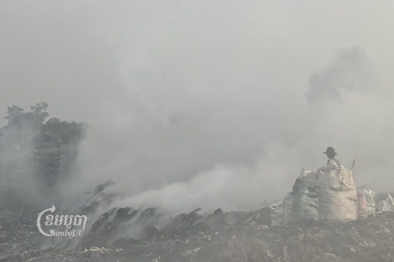 ស្រ្តីម្នាក់ឈរក្បែបាវអេតចាយដែលបានប្រមូលពីទីលានចាក់សំរាម ដែលពេញទៅដោយផ្សែងខ្មៅងងឹត ហុយចេញពីគំនរសំរាមចាស់ៗ ស្ថិតនៅភូមិអូរមុនី ឃុំអន្លង់វិល ស្រុកសង្កែ ខេត្តបាត់ដំបង។ រូបថតនៅថ្ងៃទី១១ ខែមីនា ឆ្នាំ២០២៣។ ខេមបូចា/ សុវណ្ណ ស្រីពេជ្រ