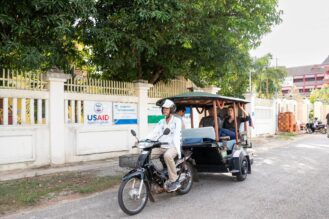 លោកស្រី សាម៉ាន់ថា ផាវើ នាយិកាទីភ្នាក់ងារ​សហរដ្ឋអាមេរិក​សម្រាប់ការអភិវឌ្ឍអន្តរជាតិ​ ហៅកាត់ថា USAID ជិះរ៉ឺម៉កកង់បីកាត់មុខ​ការិយាល័យដែលមានស្លាកសញ្ញា USAID នៅខេត្តសៀមរាប អំឡុងពេលលោកស្រីមកទស្សនកិច្ច​នៅ​កម្ពុជាក្នុងខែតុលា ឆ្នាំ២០២៤។ (រូបភាព​៖ USAID Cambodia)