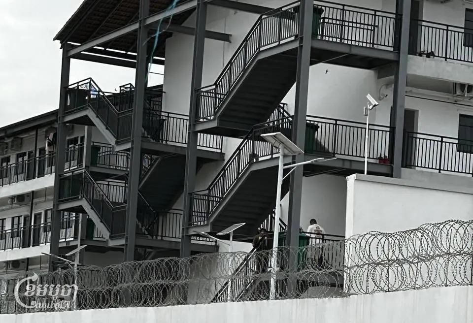 មនុស្សនៅក្នុងបរិវេណដែលហ៊ុំព័ទ្ធដោយរបងបន្លាលួសដឺរឹងមាំ ក្នុងខេត្តតាកែវ ដែលគេសង្ស័យថា មានទំនាក់ទំនងទៅនឹងទីតាំងមួយក្បែរនោះ ដែលត្រូវបាននគរបាលបង្រ្កាបកាលពីខែតុលា ក្រោយមានសេចក្តីរាយការណ៍អំពីការធ្វើទារុណកម្មនៅទីនោះ។ រូបថតថ្ងៃទី ៤ ខែធ្នូ ឆ្នាំ ២០២៤។ (ខេមបូចា/ Coby Hobbs)